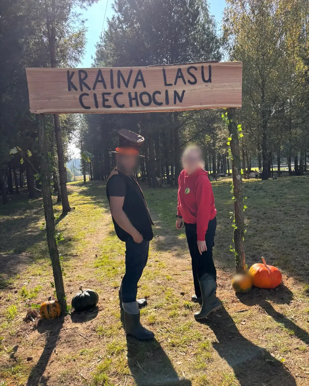 Drewniany szyld Kraina Lasu Ciechocin nad leśną drogą, pod nim dwie osoby i dynie