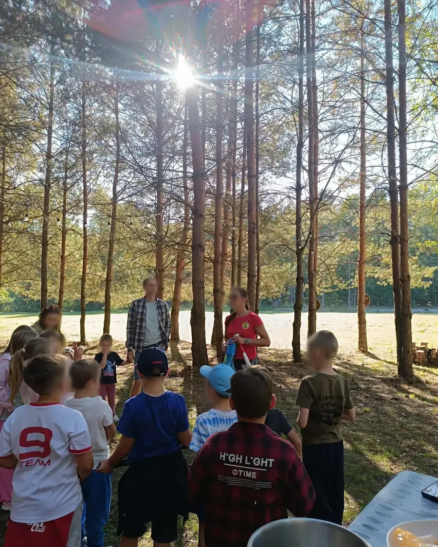 Grupa dzieci z animatorami między sosnami, przez gałęzie świeci słońce